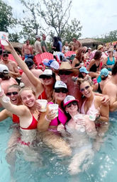Smiling group of friends in bikinis cheering with plastic cups in a crowded outdoor pool party, red lounge chairs and trees in the background on a sunny summer day.