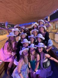 Group of young women smiling in white cowboy hats on a wooden party wagon with blue string lights and a downtown city skyline at night.