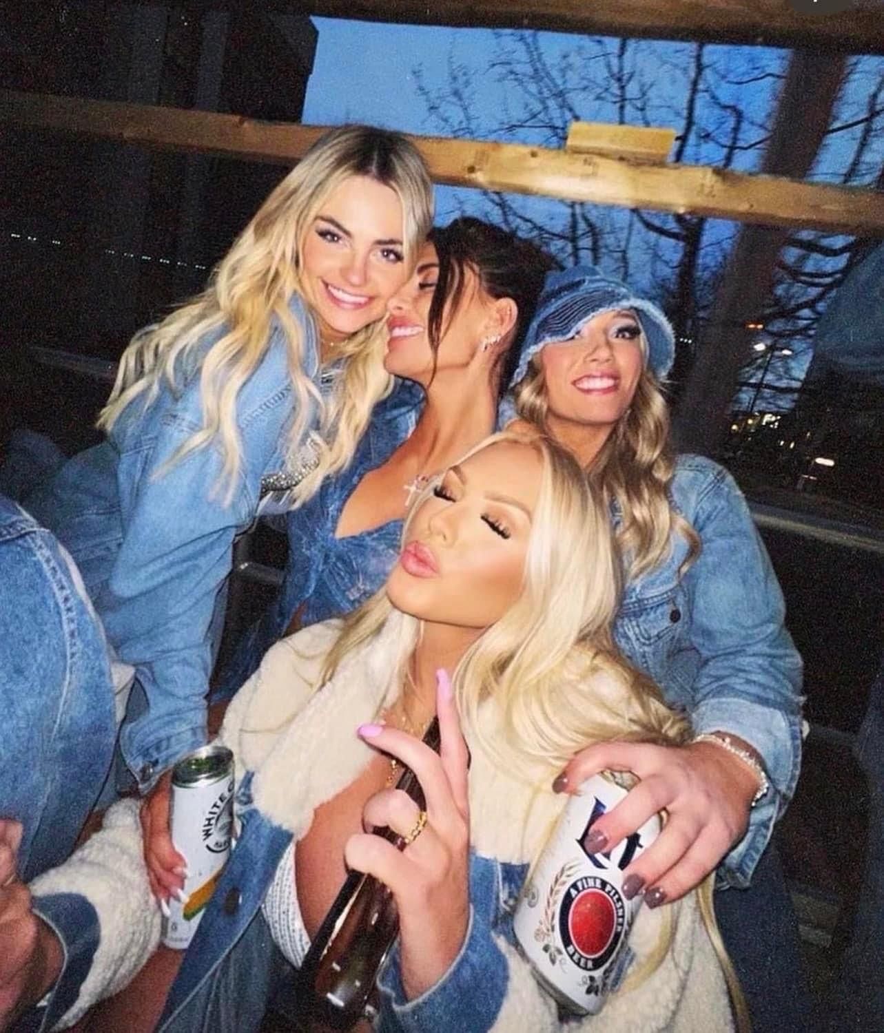 Four women in denim jackets and a bucket hat laughing and posing closely on an outdoor deck at dusk while holding beer cans and a bottle — friends’ night out.