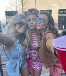 Three friends taking a playful daytime selfie on a downtown urban street — center in a pink zebra dress with a tinsel crown, left in a light-blue fringed romper and sunglasses, right in a brown jacket and sunglasses reaching forward with a red party cup.