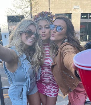 Three friends taking a playful daytime selfie on a downtown urban street — center in a pink zebra dress with a tinsel crown, left in a light-blue fringed romper and sunglasses, right in a brown jacket and sunglasses reaching forward with a red party cup.