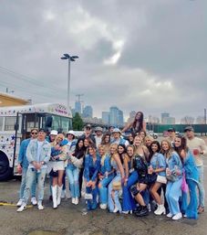 Large group of friends in denim and festival outfits posing by a star‑decorated party bus in a downtown parking lot under cloudy skies, upbeat urban party vibe