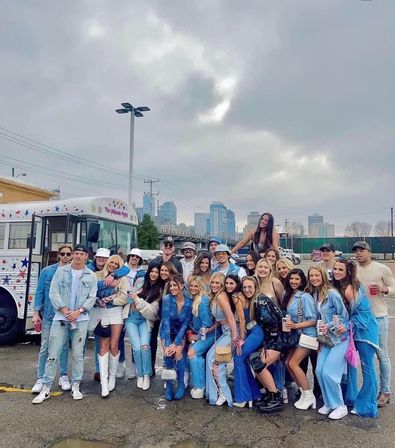 Large group of friends in denim and festival outfits posing by a star‑decorated party bus in a downtown parking lot under cloudy skies, upbeat urban party vibe
