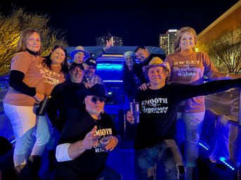 Cheerful group of adults at a nighttime rooftop deck party with blue LED lights and city skyline in the background, wearing casual tees and cowboy hats, holding drinks and smiling for the camera.