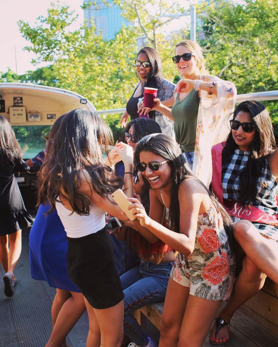 Group of friends enjoying a sunny outdoor boat party — women in summer outfits and sunglasses laughing, taking selfies and holding drinks with trees and city buildings behind them.