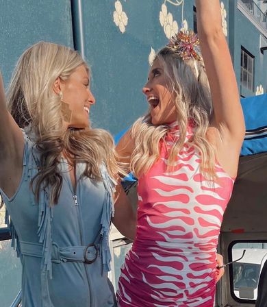 Two women cheering outdoors by a blue floral mural — one in a pink zebra-print dress and sparkly crown, the other in a light-blue fringed romper.