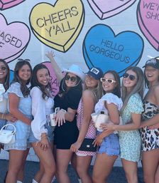 Group of friends in summer outfits laughing and posing in front of a colorful candy-heart mural that reads 'CHEERS Y'ALL' and 'HELLO GORGEOUS'.