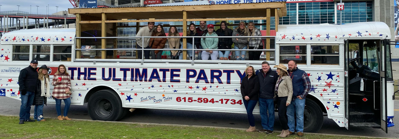 Smiling group of friends posing on and beside a white, star-spangled party bus with an open upper deck, parked outside a sports stadium for a lively outdoor tailgate celebration.