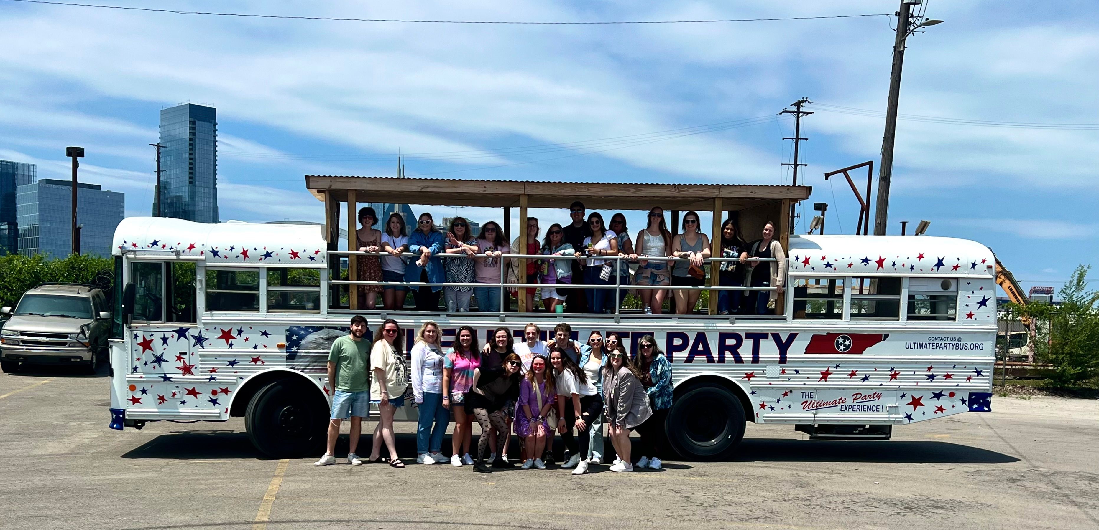Group of friends posing with an open-air, star-spangled party bus with an upper deck in a downtown parking lot under a bright blue summer sky