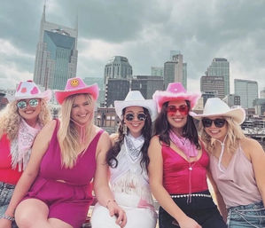 Five friends in pink and white cowgirl hats, sunglasses and playful party outfits sitting on a rooftop with a modern downtown skyline under a cloudy sky