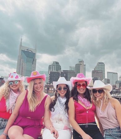 Five smiling friends in pink outfits and cowboy hats posing on a rooftop with the Nashville skyline and cloudy sky behind them.