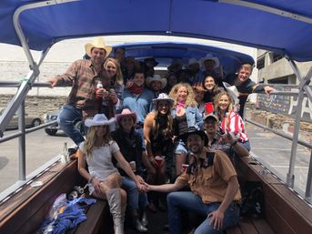 Daytime group of friends on a covered party trolley in a downtown parking lot, wearing cowboy hats, denim and boots, holding red cups and beers.