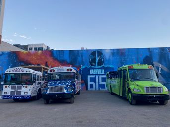 Three colorful party buses (white with stars, blue with flag pattern, and bright green) parked in an open lot in front of a large Nashville mural featuring a football helmet and the number 615 under a clear blue sky.