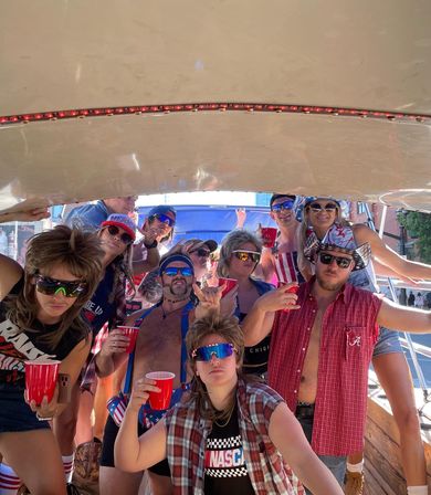 Group of friends at a summertime boat party wearing patriotic red, white and blue outfits, mullet wigs and sunglasses, holding red solo cups and striking playful poses under a boat canopy.
