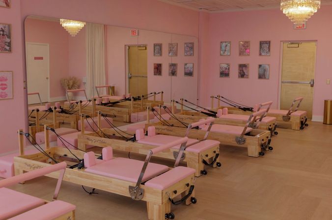 Boutique pink Pilates studio interior with rows of wooden reformer machines, mirrored wall, chandeliers, and light wood floors.