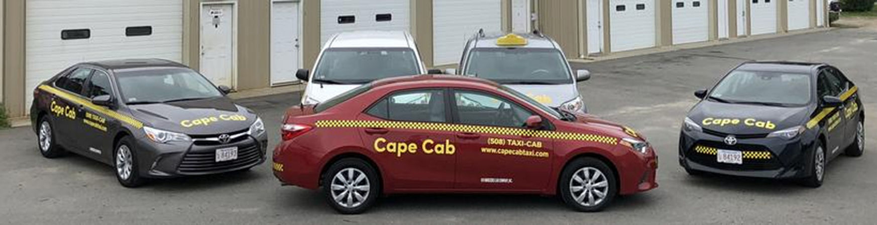 Fleet of four taxis parked outside garage bays on an asphalt lot: red compact sedan with yellow checker stripe, two dark gray sedans and a white taxi minivan with roof sign