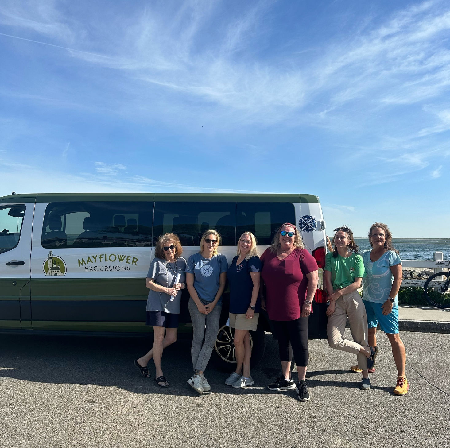 Six women smiling and posing beside a green tour van parked on an oceanfront road under a bright blue sky, ready for a coastal excursion.