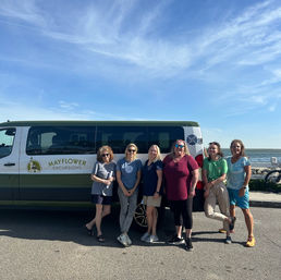 Six women smiling and posing beside a green tour van parked on an oceanfront road under a bright blue sky, ready for a coastal excursion.