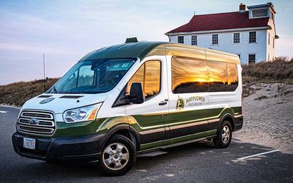 White-and-green passenger shuttle van parked on a coastal road by sand dunes and a clapboard beach house, golden-hour sunset reflecting in its windows.
