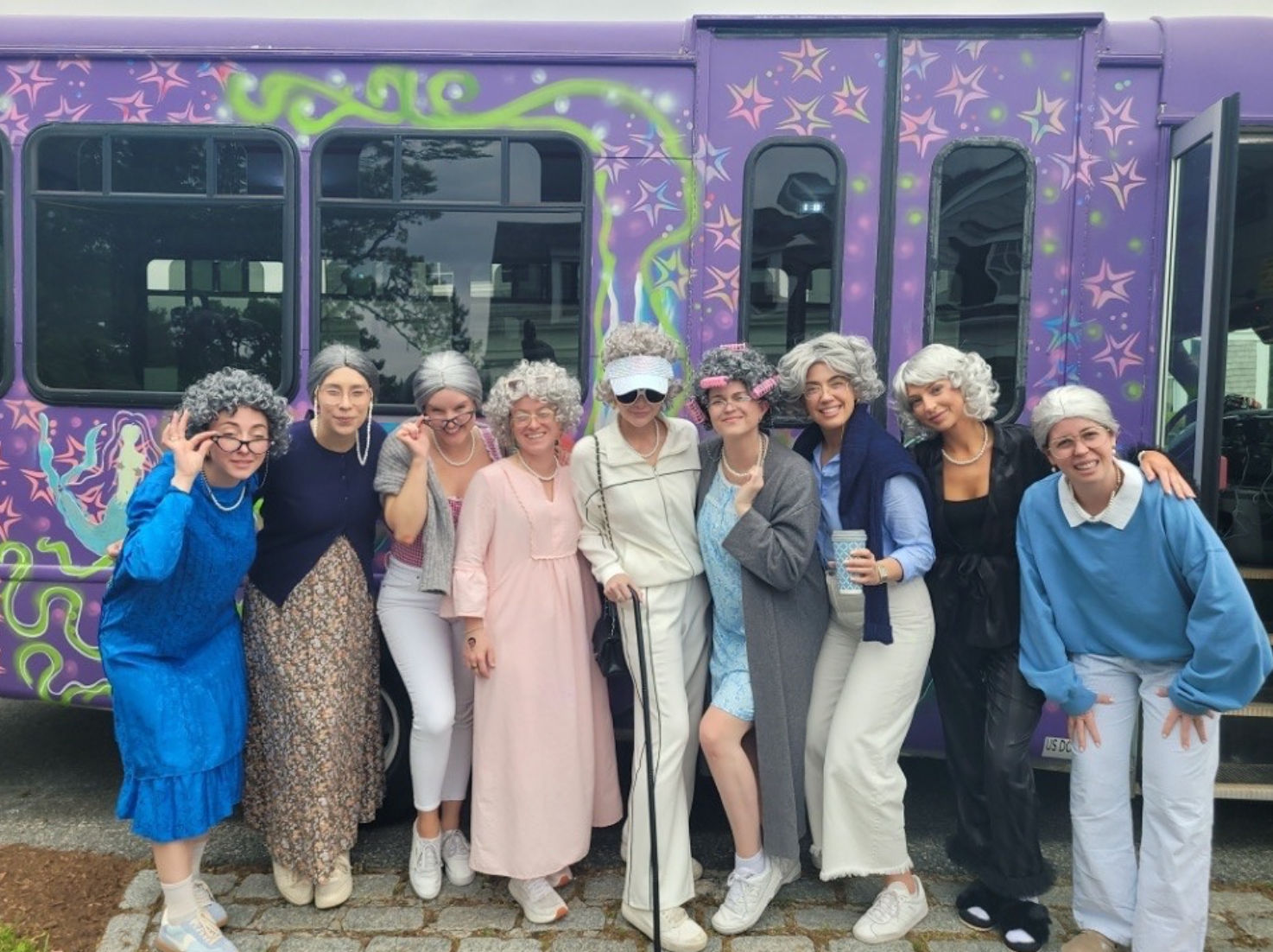 Nine people in gray wigs and playful vintage 'grandma' costumes smiling and posing on a cobblestone curb in front of a purple, star-patterned party bus