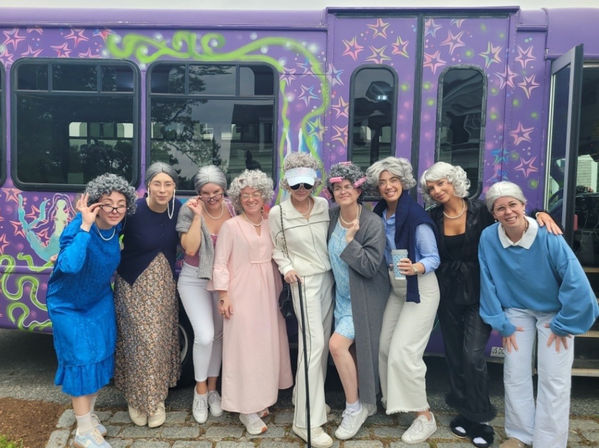 Nine people in gray wigs and playful vintage 'grandma' costumes smiling and posing on a cobblestone curb in front of a purple, star-patterned party bus