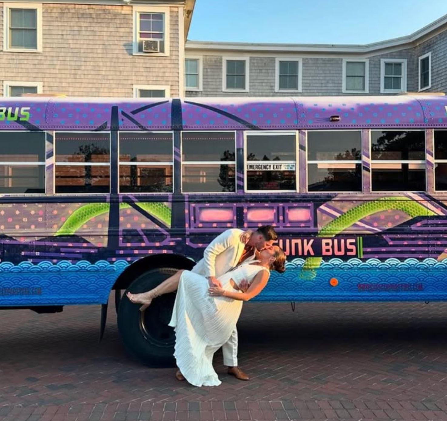 Newlywed couple in wedding attire share a dip-and-kiss in front of a colorful purple party bus with wave graphics, parked by shingled seaside buildings at sunset.