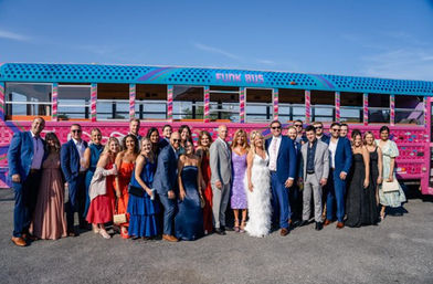 Large group of well-dressed wedding guests and friends posing cheerfully in front of a vibrant pink-and-blue party bus under a clear blue sky.