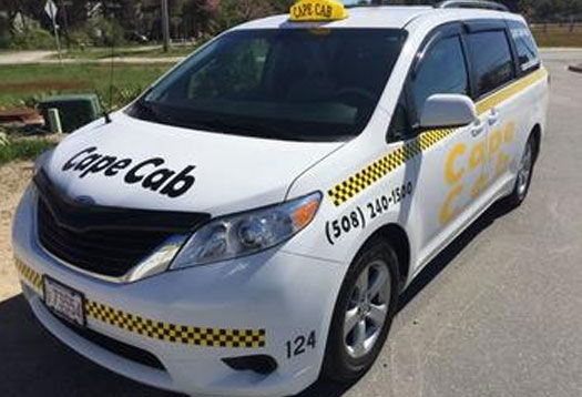 White taxi minivan with yellow roof sign and black-and-yellow checker stripe parked curbside on a sunny suburban street, ready to pick up passengers