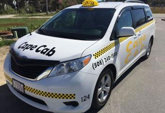 White taxi minivan with yellow roof sign and black-and-yellow checker stripe parked curbside on a sunny suburban street, ready to pick up passengers
