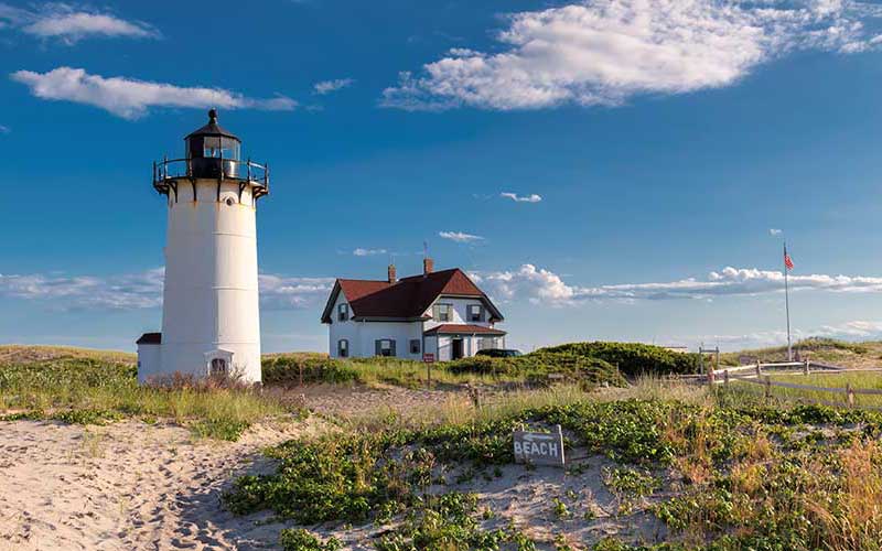 White lighthouse beside a classic keeper's house on grassy sand dunes, a small beach sign in the foreground and an American flag under a bright blue sky