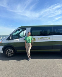 Person in a green shirt leaning against a green-and-white coastal tour van parked at a sunny waterfront with calm sea and blue sky.