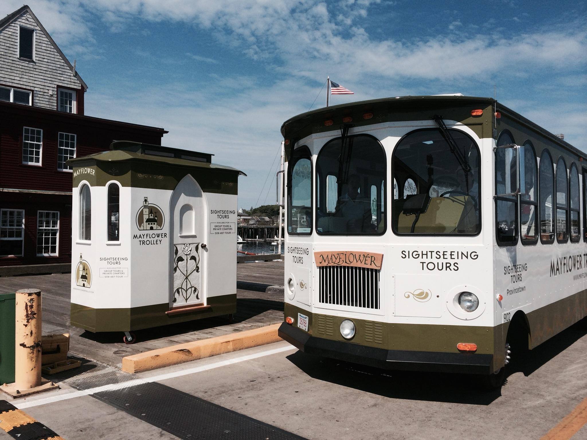 Vintage-style white and olive-green sightseeing trolley parked at a sunny coastal pier beside a matching ticket kiosk, blue sky and American flag in the background.