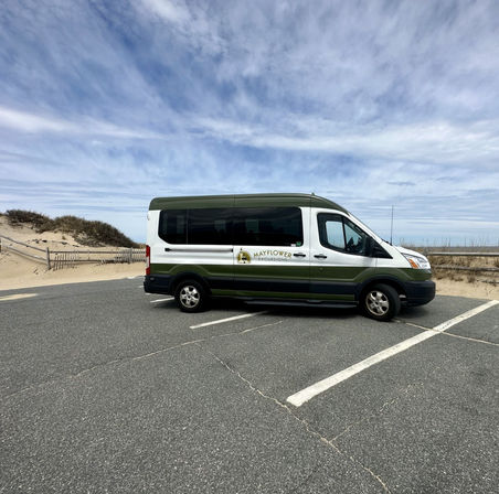 Green-and-white beach shuttle van parked in a coastal parking lot beside sand dunes and beach grasses under a wide cloudy blue sky