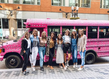 Eight women posing and smiling on a downtown city sidewalk in front of a bright pink leopard-print party bus, casual outfits and urban buildings in the background.
