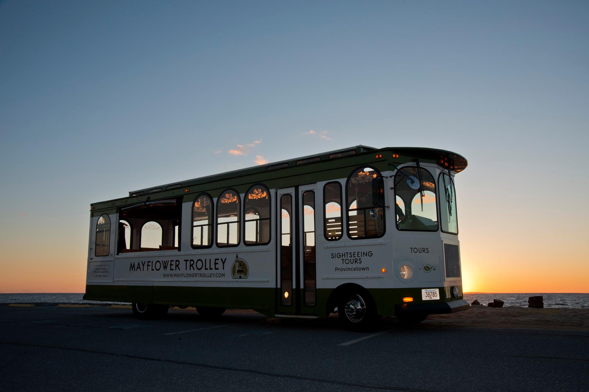 Coastal green-and-white vintage sightseeing trolley parked on an oceanfront road at sunset, silhouetted against a colorful sky with warm reflections on its windows.