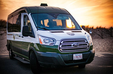 White-green coastal shuttle van parked by sandy beach dunes at sunset, glowing under a golden sky