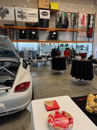 White sports car in an indoor automotive warehouse hosting a holiday gathering with high shelving and parts banners, cocktail tables with poinsettias, and a table with "Merry Christmas" plates and sandwiches.