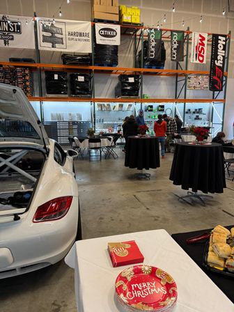 White sports car in an indoor automotive warehouse hosting a holiday gathering with high shelving and parts banners, cocktail tables with poinsettias, and a table with "Merry Christmas" plates and sandwiches.