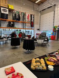 Catered holiday event in an industrial warehouse: sandwich and charcuterie buffet, high-top tables with poinsettias, string lights, open garage door with a car display and inflatable play structure, shelving and boxes in the background.