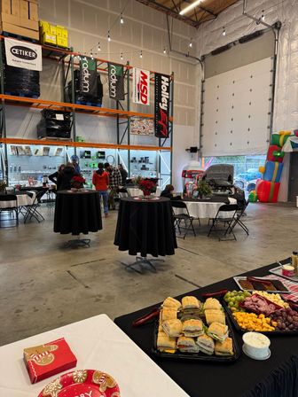 Catered holiday event in an industrial warehouse: sandwich and charcuterie buffet, high-top tables with poinsettias, string lights, open garage door with a car display and inflatable play structure, shelving and boxes in the background.