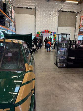 Green and gold vintage race car parked inside a warehouse car show with a colorful 'Merry Christmas' inflatable arch, display cases of model cars, merch racks and visitors under string lights.
