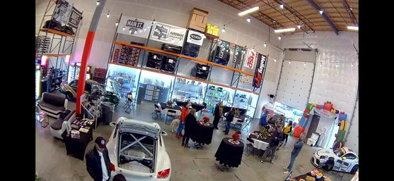 Bright warehouse-style auto shop showroom with race cars on display, open shelving, folding tables and people mingling under high ceilings