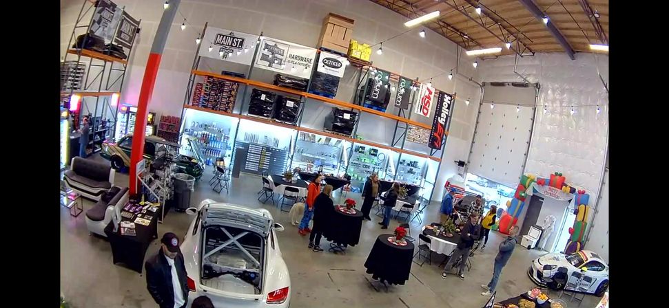 Bright warehouse-style auto shop showroom with race cars on display, open shelving, folding tables and people mingling under high ceilings
