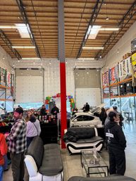 Busy indoor car meet in a bright warehouse garage — sofas and a display table in the foreground, people mingling around sport cars with open hoods, exposed wooden ceiling beams, colorful banners and a holiday inflatable arch in the back.