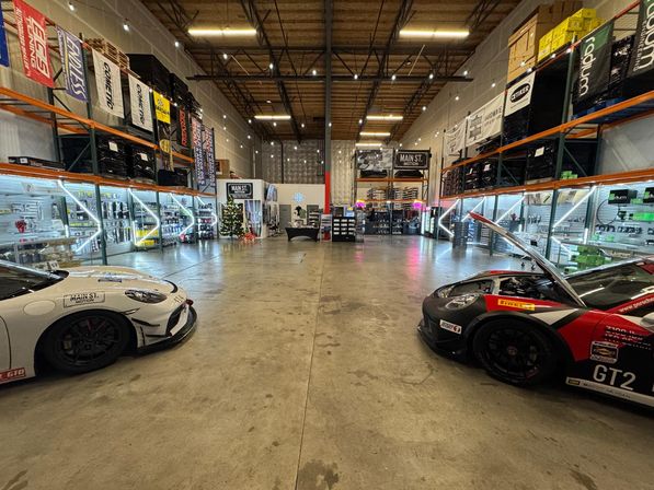 Wide warehouse-style auto performance shop showroom with two GT race cars parked nose-in on either side of a central concrete aisle, illuminated display racks of performance parts, tall shelving and banners overhead, and a small Christmas tree near the back.