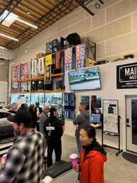 Lively indoor motorsports garage meetup with people chatting around a displayed car and couches, tall warehouse shelving lined with racing banners, a wall-mounted TV showing a race, monitors and a whiteboard under a high ceiling.