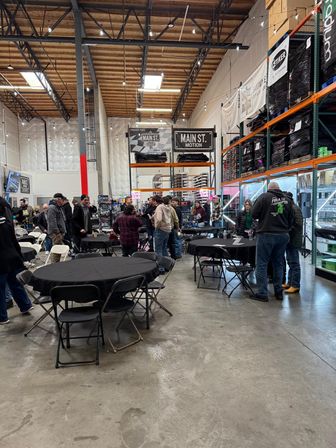 Bustling indoor warehouse meet-up with round tables covered in black cloth, folding chairs, people mingling beside tall industrial shelving stocked with boxed products under exposed wooden ceiling beams.