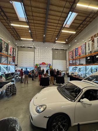 White Porsche in foreground at an indoor car meet inside a high-ceiling warehouse garage with skylights and string lights, people gathered around tall tables, and automotive banners and parts shelving along the walls.