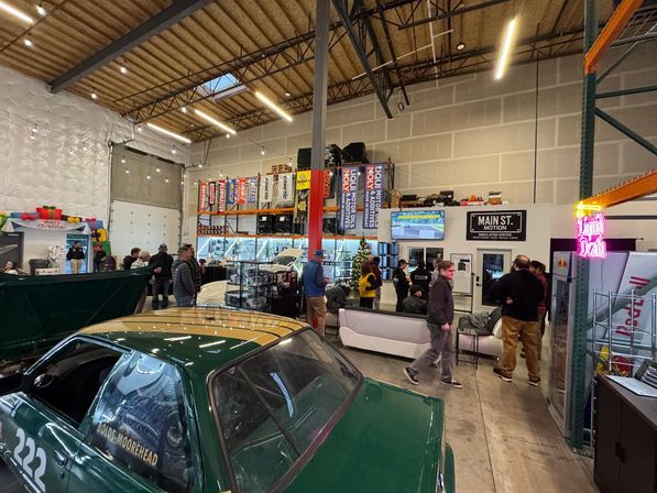 Warehouse-style auto shop showroom event with a green race car in the foreground, people browsing displays and sitting on a white couch, tall shelving with automotive banners, neon sign and holiday decorations.