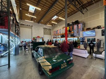 Green and gold BMW race car #222 parked in a bright warehouse garage during an indoor car meet — people mingling, service bays, tall shelving, banners, and TV screens.